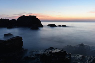 Kıyı boyunca güneş ufukta batarken kayalar ve deniz dalgaları vardır. Karanlık kayalar var çünkü çok az ışık var. Ve yazın denizin yumuşak dalgaları