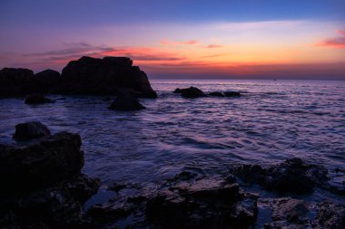 Kıyı boyunca güneş ufukta batarken kayalar ve deniz dalgaları vardır. Karanlık kayalar var çünkü çok az ışık var. Ve yazın denizin yumuşak dalgaları