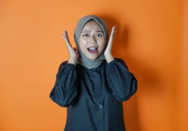 Bir tesettür takan Asyalı kadın portresi, iki eli yanaklarının yanında sürpriz bir poz veriyor. Turuncu arka planda izole edilmiş..