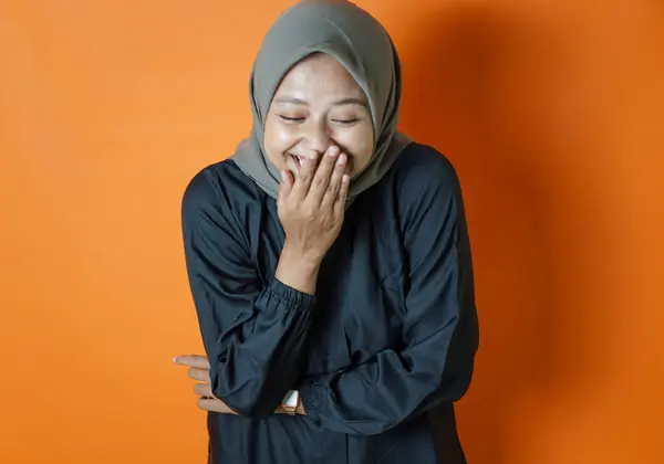 Başörtüsü takan mutlu bir Asyalı kadının portresi. Ağzını kapatırken gülümsüyormuş gibi poz veriyor. Turuncu arka planda izole edilmiş..