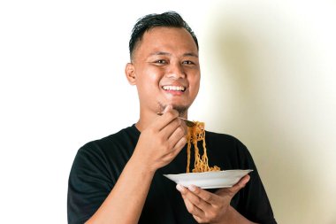 Siyah gömlek giyen bir adam bir tabak erişte tutuyor ve çatalla yemek yiyor. Memnun görünüyor ve beyaz bir arkaplanda poz veriyor.