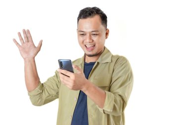 Bir adam akıllı telefonuna bakarken gülümsüyor, bir elini kaldırarak selamlıyor. Bir video görüşmesinden ya da dijital bir etkileşimden hoşlanıyor gibi görünüyor..