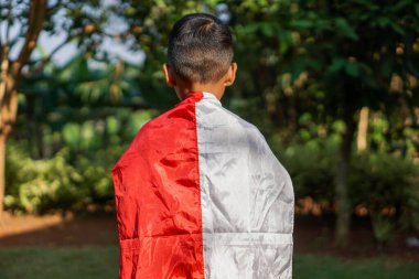 Arkadan bir çocuk görülüyor, Endonezya bayrağına sarılmış, yeşil ağaçlardan ve bitkilerden oluşan arka planda duruyor, ulusal gururu simgeliyor..