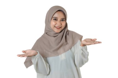 Bej tesettürlü ve beyaz bluzlu bir kadın, iki eli havada, avuçları havada, düz beyaz bir arka plana doğru hareket ederken gülümsüyor..
