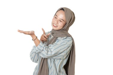 Çizgili gömlekli ve kahverengi tesettürlü bir kadın gülümseyip açık elini göstererek beyaz bir arka plan karşısında bir şey ima ediyor ya da bir fikir sunuyor..