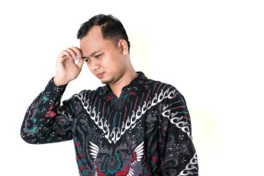 Renkli batik tişörtlü bir adam derin düşüncelere dalmış beyaz bir arka plana karşı duruyor. Bir eli alnında, dalgın ve odaklanmış görünüyor. Endonezya Batik Günü