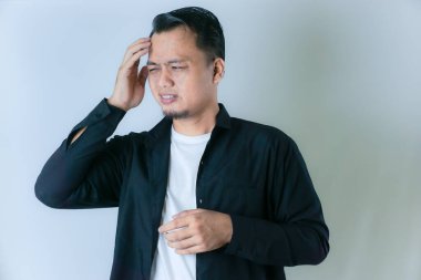 Siyah gömlekli ve beyaz tişörtlü bir adam başını tutuyor, acı çekiyor ya da acı çekiyor gibi görünüyor, düz bir arka planda..