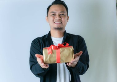 Siyah tişörtlü ve beyaz tişörtlü bir adam kırmızı kurdeleyle sarılı altın bir hediye kutusunu beyaz arka planda sunarken gülümsüyor..