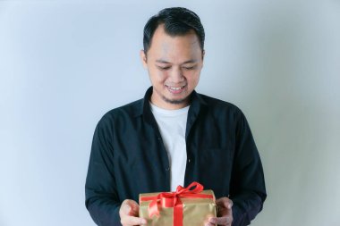 Siyah tişörtlü ve beyaz tişörtlü bir adam kırmızı kurdeleyle sarılı altın bir hediye kutusunu beyaz arka planda tutarken gülümsüyor..