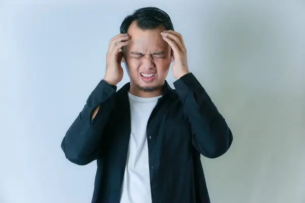 Siyah gömlekli ve beyaz tişörtlü bir adam başını tutuyor, acı çekiyor ya da acı çekiyor gibi görünüyor, düz bir arka planda..