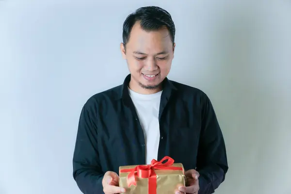 Siyah tişörtlü ve beyaz tişörtlü bir adam kırmızı kurdeleyle sarılı altın bir hediye kutusunu beyaz arka planda tutarken gülümsüyor..