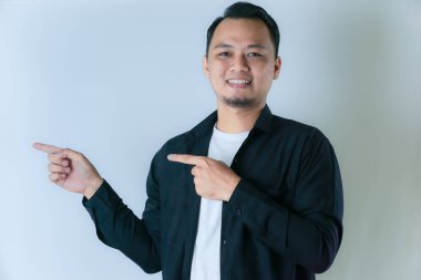 Siyah tişörtlü, beyaz tişörtlü ve kısa saçlı bir adam gülümsüyor ve iki eliyle solu işaret ediyor..