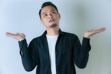 Siyah tişörtlü ve beyaz tişörtlü bir adam, beyaz bir arka plana karşı sorgulama ifadesiyle iki elini kaldırıyor gibi görünüyor..