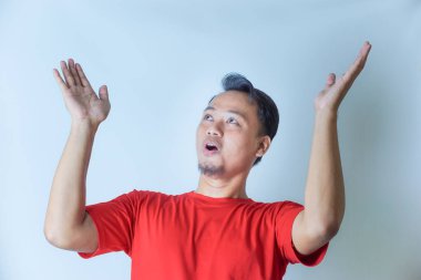 Kırmızı gömlekli bir adam sevinçle kollarını kaldırır yukarı bakar, heyecanını ifade eder ya da sade bir arka plana karşı kutlama yapar..