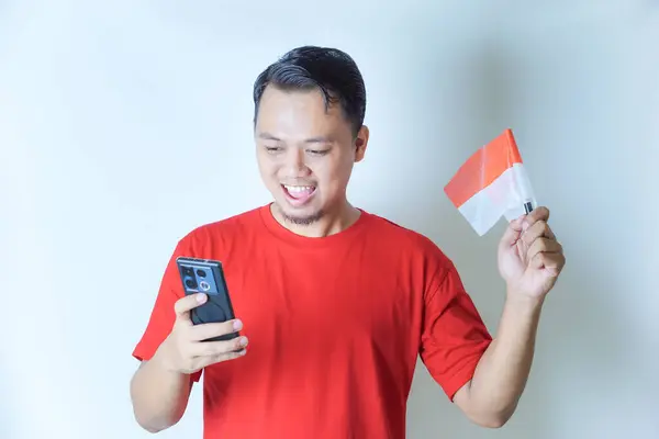 Kırmızı gömlekli bir adam telefonuna bakıp gülümseyerek Endonezya bayrağını tutarken Endonezya 'nın bağımsızlık gününü kutluyor..