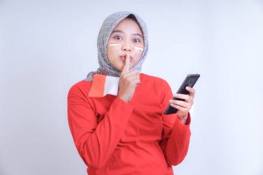 Kırmızı yüzlü, bayrak boyalı ve bayrak eşarplı bir kadın susma hareketi yapıyor, elinde telefon var. Endonezya Bağımsızlık Günü kutlaması kavramı.