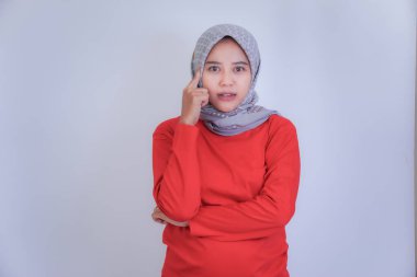 Kırmızı bluzlu ve gri tesettürlü bir kadın eli çenesinde beyaz bir arka plana karşı düşünceli bir şekilde poz veriyor..