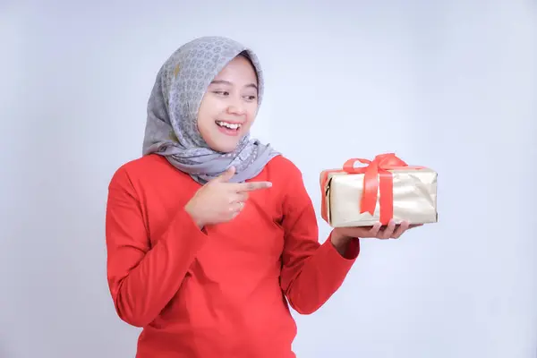 Kırmızı tişörtlü ve tesettürlü bir kadın gülümsüyor ve elinde kırmızı kurdeleli altın bir kağıda sarılı bir hediye taşıyor..