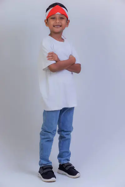 Beyaz gömlekli, mavi kotlu ve kırmızı-beyaz bantlı genç bir çocuk kollarını kavuşturmuş duruyor..