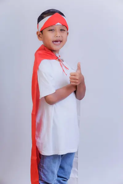 stock image A boy wearing a red and white headband and Indonesian flag on his back cape gives a thumbs-up, smiling confidently.