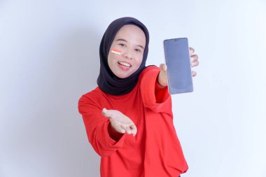 Endonezyalı genç bir kadın, beyaz arka planda izole edilmiş, boş ekran akıllı telefonu tutarken ve sunarken yanağında Endonezya bayrağı olan kırmızı bir gömlek giyiyor.