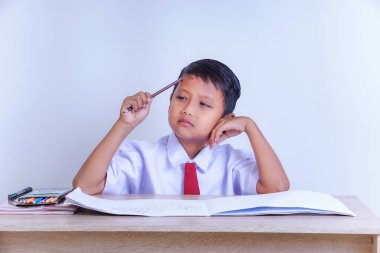 Okul üniformalı Endonezyalı genç bir çocuk kafasında kalemle düşünceli görünüyor. Masasında açık bir defter ve renklerle oturuyor..