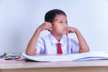 Okul üniformalı Endonezyalı genç bir çocuk kafasında kalemle düşünceli görünüyor. Masasında açık bir defter ve renklerle oturuyor..