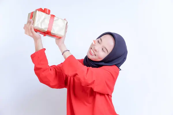 Endonezyalı genç bir kadın tesettür takıyor, yanağında küçük bir bayrak etiketi var, kırmızı kurdeleye sarılı bir hediye verirken gülümsüyor, Endonezya 'nın bağımsızlık gününü 17 Ağustos' ta kutluyor..