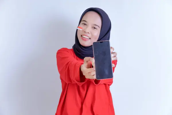 Endonezyalı genç bir kadın tesettürlü ve kırmızı bluzlu cep telefonunu gösterirken gülümsüyor. Yanağı beyaz arka planda izole edilmiş Endonezya bayrağı çıkartmasıyla süslenmiş.