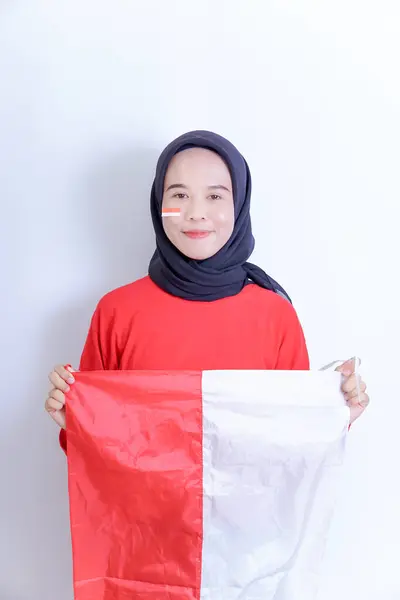 Genç Endonezyalı tesettürlü bir kadın kırmızı ve beyaz bir bayrak tutuyor, yanaklarında aynı renk çizgiler var. Gülümser ve doğrudan kameraya bakar, beyaz bir arkaplanda izole bir şekilde..