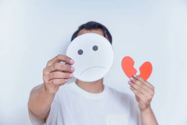 stock image A person expresses sadness by holding a frowning face mask and a broken heart symbol, symbolizing emotional pain. world mental health day concept