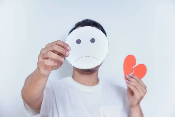 stock image A person expresses sadness by holding a frowning face mask and a broken heart symbol, symbolizing emotional pain. world mental health day concept