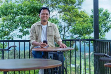 Neşeli Asyalı bir adam açık havada, yemyeşil ağaçların arasında, ahşap bir masada oturmaktan zevk alıyor..