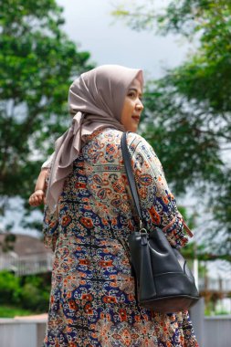 Genç bir Asyalı Müslüman anne desenli bir elbise ve tesettür içinde çocuğunu yemyeşil bir ortamda taşıyor..