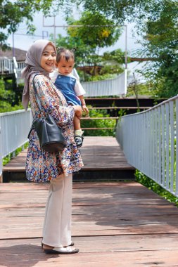 Mutlu, genç bir Asyalı anne, yeşilliklerle çevrili ahşap bir köprüde yürürken bir çocuk taşıyor..
