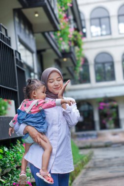 Neşeli, tesettürlü Asyalı bir kadın çiçeklerle çevrili, güzel bir doğa ortamında bir çocuğu kucaklıyor..