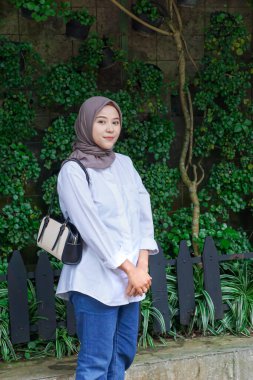 Beyaz gömlekli ve tesettürlü genç bir Asyalı kadın canlı bitkilerle yeşil bir ortamda poz veriyor..