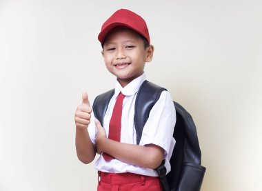 Okul üniformalı neşeli Asyalı bir çocuk sırt çantasıyla başparmağını kaldırıyor..
