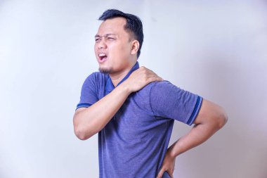 Asyalı bir adam rahatsızlık yaşıyor, omzunu tutuyor ve acı belirtileri gösteriyor..