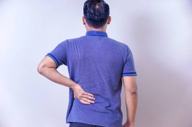 Mavi tişörtlü Asyalı bir adam belini tutuyor, muhtemelen rahatsız olduğunu gösteriyor..