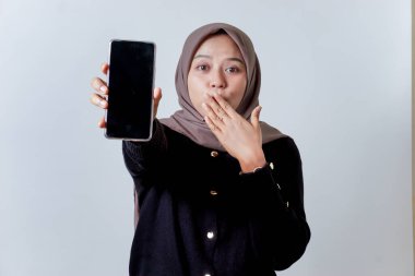 Başörtüsü takan Asyalı genç bir kadın şakacı bir şekilde ağzını kapatırken telefonu tutuyor ve sunuyor..