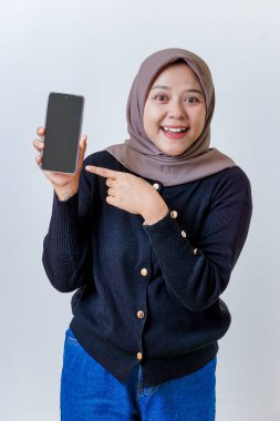 Tesettürlü gülümseyen Asyalı bir kadın akıllı telefonunu kendine güvenle işaret ederken gösterir..