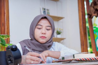 Tesettürlü Asyalı bir kadın samimi bir çalışma ortamında özenle notlar yazıyor.