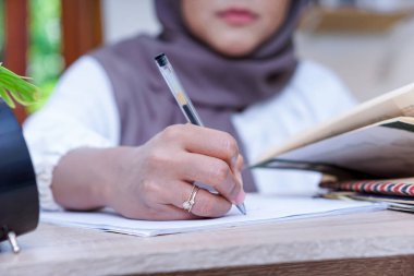 Tesettürlü bir kadın masasında kalemle notlar yazıyor. Etrafı bitki ve saatle çevrili..