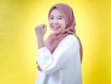 Tesettürlü neşeli bir Asyalı kadın, parlak sarı bir arka plana karşı ellerini kaldırarak kutluyor.