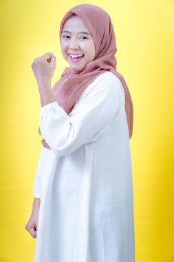 Tesettürlü neşeli bir Asyalı kadın, parlak sarı bir arka plana karşı ellerini kaldırarak kutluyor.