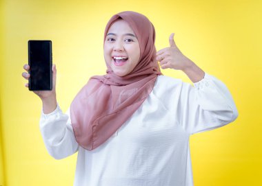 Tesettürlü neşeli Asyalı bir kadın akıllı bir telefon tutar ve parlak sarı bir arka plana başparmak atar..