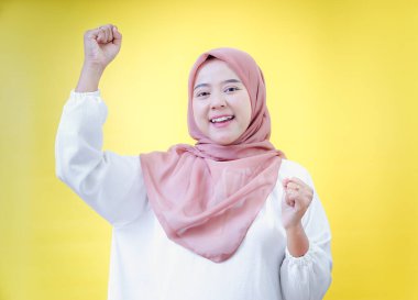 Mutlu Asyalı kadın tesettürlü, açık sarı bir arka planda yumruğunu kaldırarak kutluyor.
