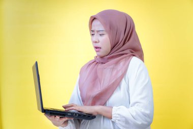 Asyalı bir kadın, dizüstü bilgisayarını parlak sarı bir arka plan önünde kullanırken şaşkın görünür..