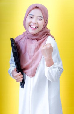 Beyaz elbiseli ve tesettürlü neşeli Asyalı bir kadın dizüstü bilgisayar tutuyor, sarı bir arka plana karşı başarıyı kutluyor..